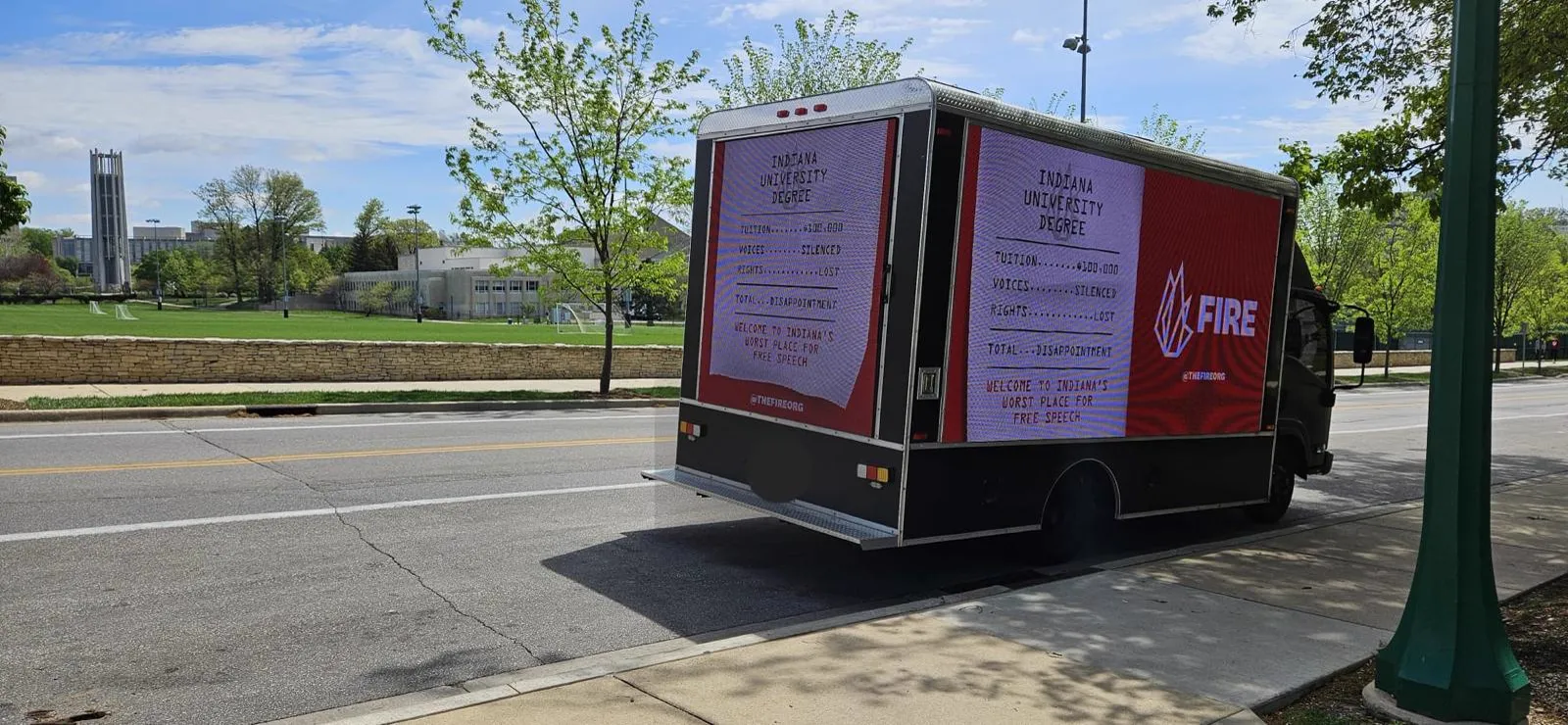 FIRE LED billboard truck parked on a public road beside Indiana University Bloomington campus green