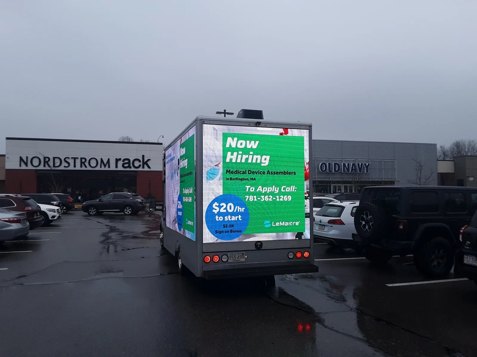 LED billboard truck parked in a shopping center lot between retail anchor stores running a Now Hiring campaign in the rain