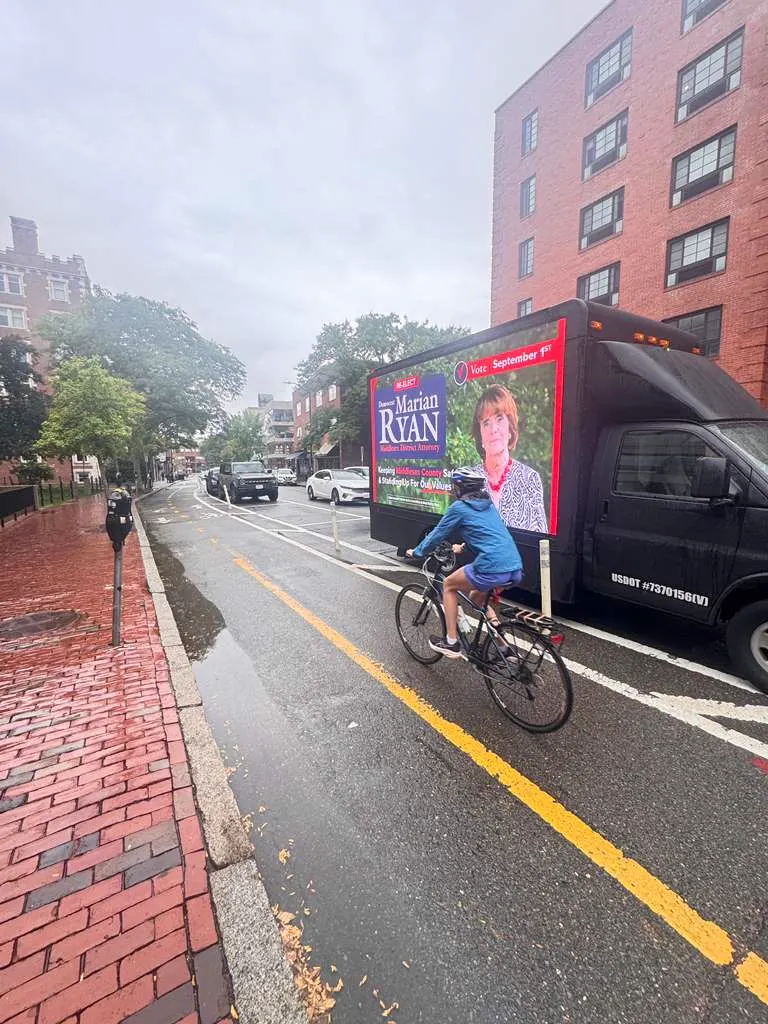 Marian Ryan campaign LED billboard truck on a rainy Cambridge street beside a cyclist in a bike lane on primary election day