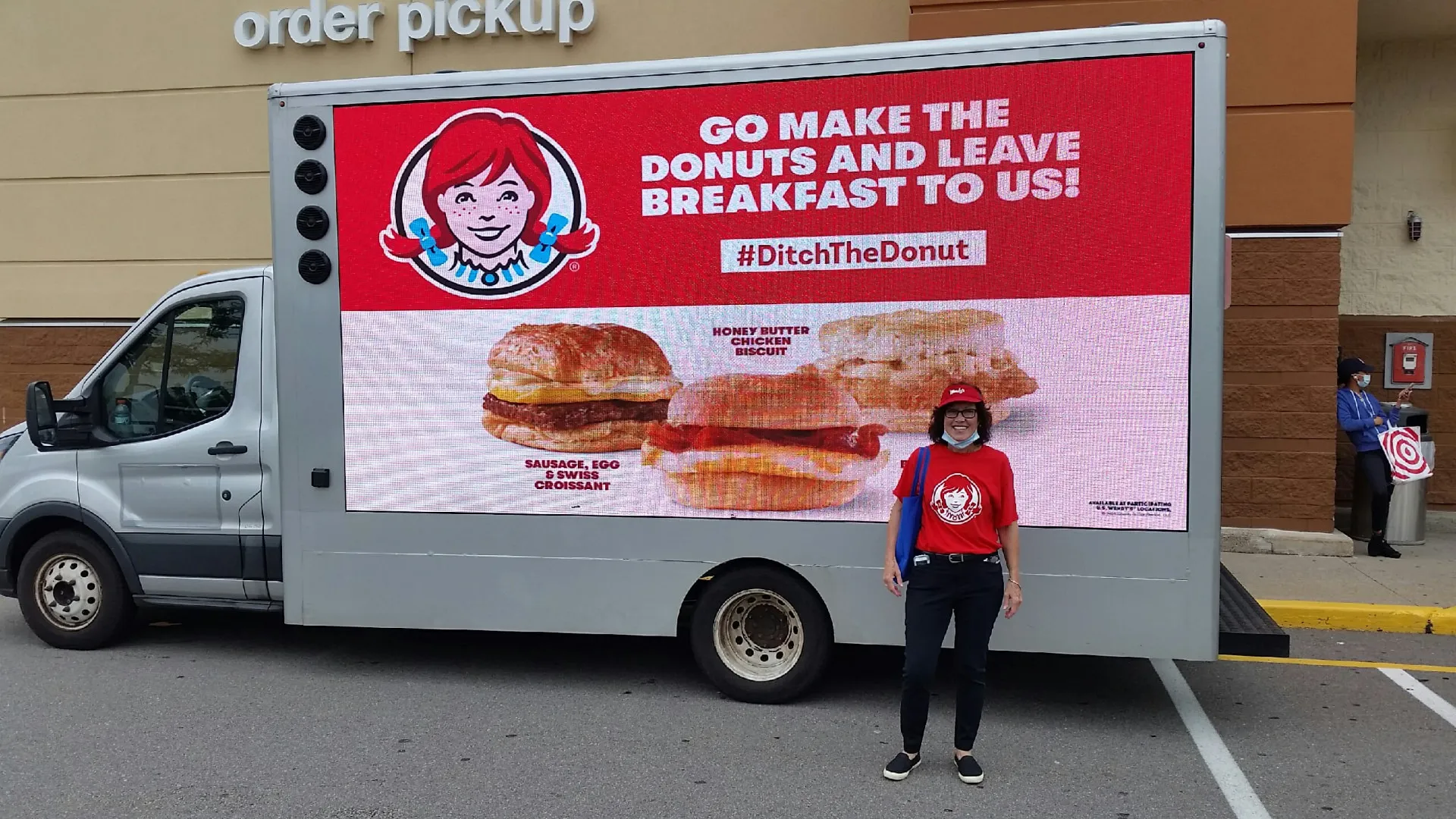 Wendy's brand ambassador standing beside the #DitchTheDonut breakfast LED billboard truck outside an order pickup entrance